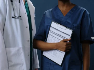 Two healthcare professionals, one in a white coat and one in dark blue scrubs, standing together while holding a medical clipboard.