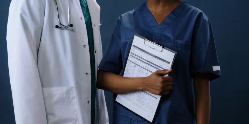 Two healthcare professionals, one in a white coat and one in dark blue scrubs, standing together while holding a medical clipboard.
