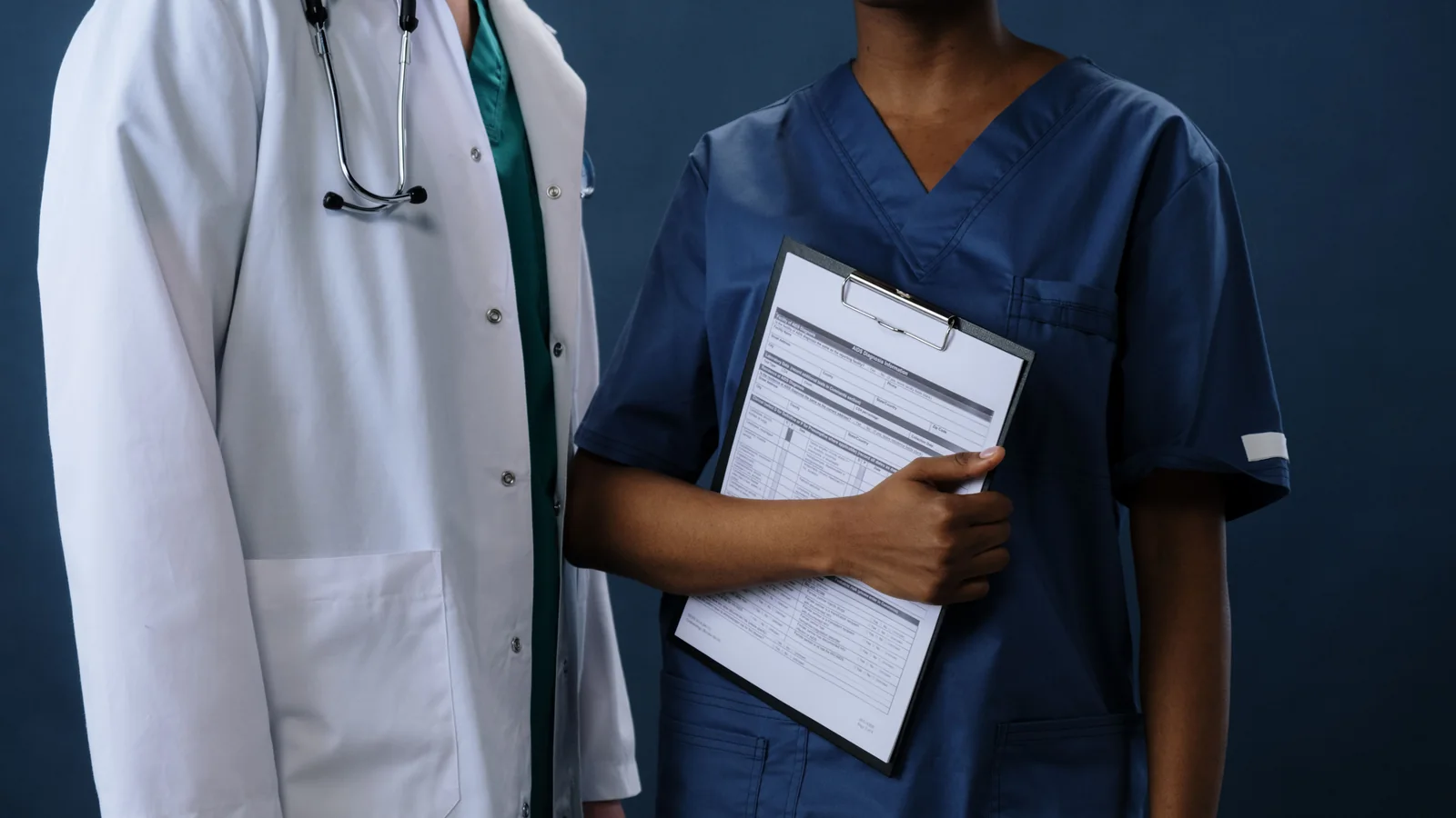 Two healthcare professionals, one in a white coat and one in dark blue scrubs, standing together while holding a medical clipboard.