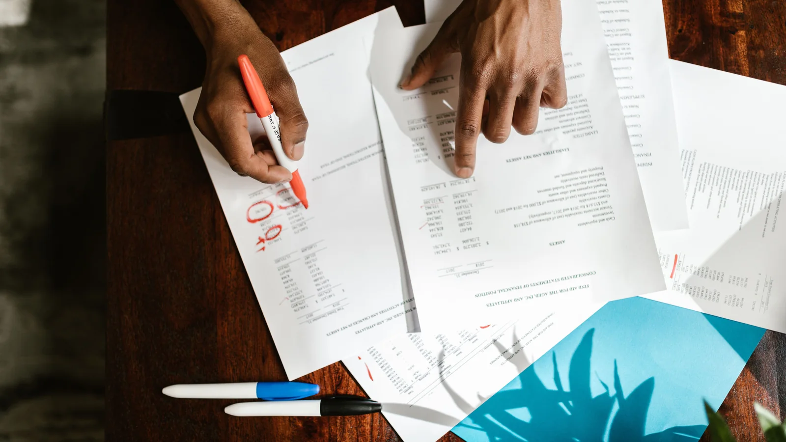 Overhead view of hands reviewing and highlighting documents spread across a wooden desk.