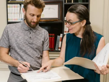 Two adults review legal documents together in an office, with one person writing while the other holds an open folder.