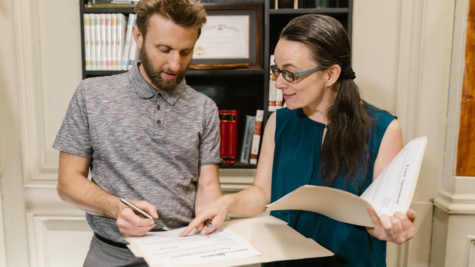 Two adults review legal documents together in an office, with one person writing while the other holds an open folder.
