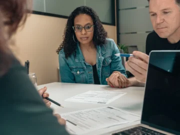 A couple reviews paperwork with a professional at a table in an office, with a laptop and documents visible.