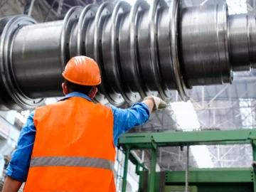 Worker in an orange hard hat and reflective vest inspects large industrial machinery inside a factory.