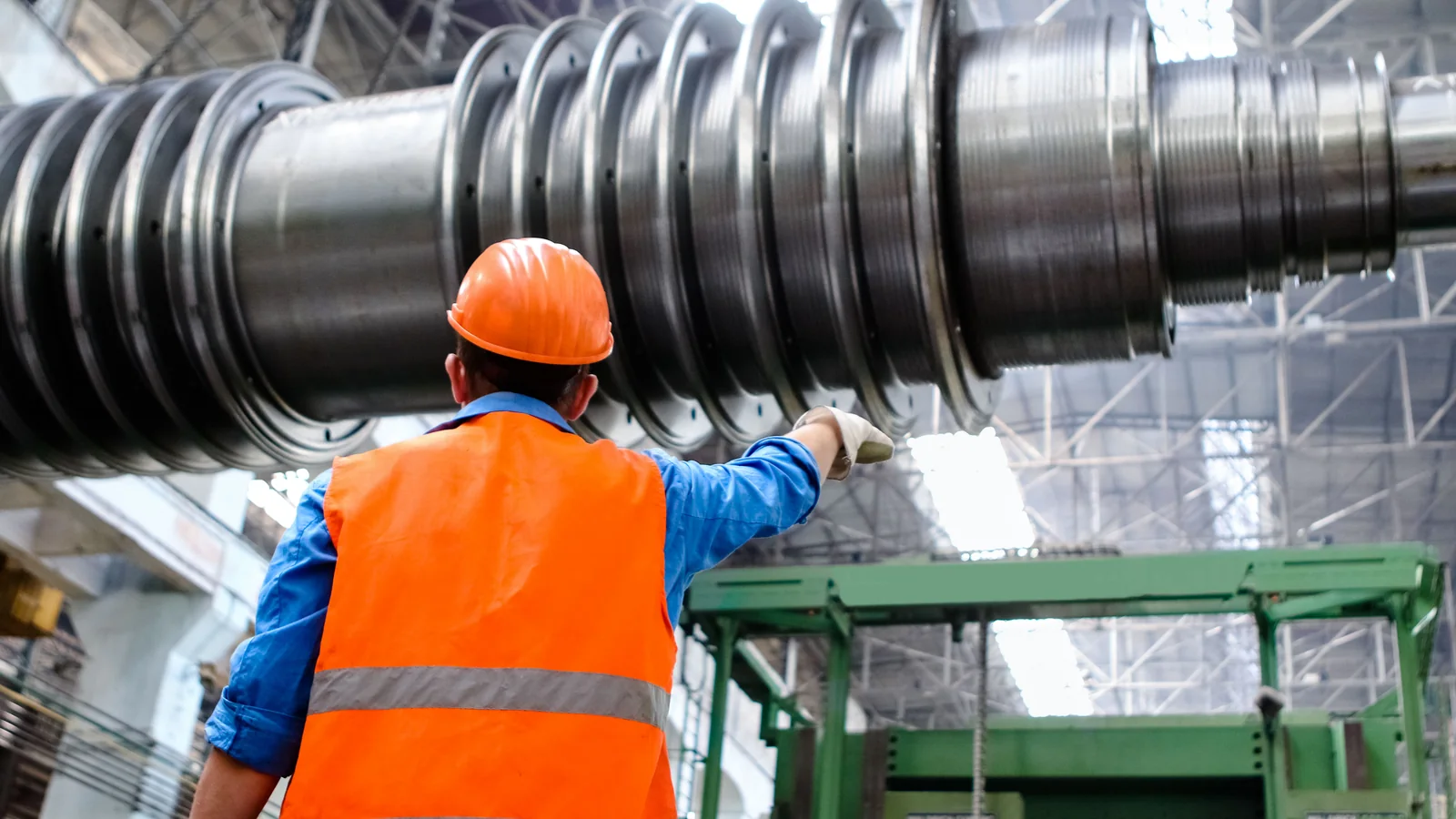 Worker in an orange hard hat and reflective vest inspects large industrial machinery inside a factory.