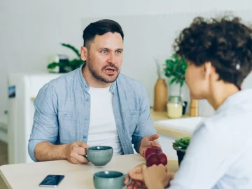 A couple having a serious conversation across a kitchen table with coffee cups.
