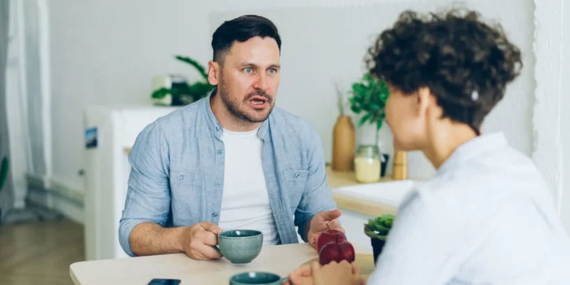 A couple having a serious conversation across a kitchen table with coffee cups.