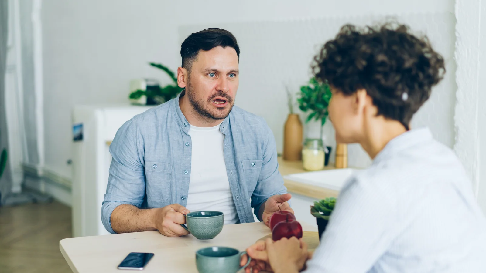 A couple having a serious conversation across a kitchen table with coffee cups.