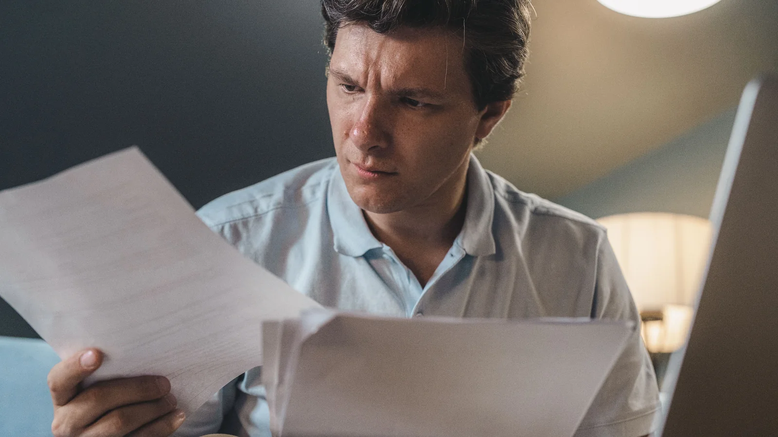 A man seated indoors carefully reviewing several sheets of paper under warm lighting.