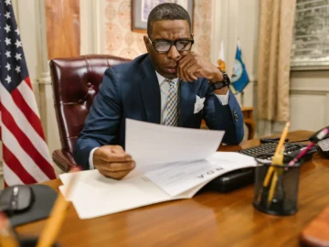 A suited professional reviews paperwork at a wooden office desk beside a U.S. flag, computer, telephone, and globe.