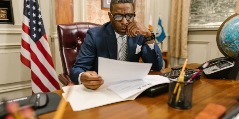 A suited professional reviews paperwork at a wooden office desk beside a U.S. flag, computer, telephone, and globe.