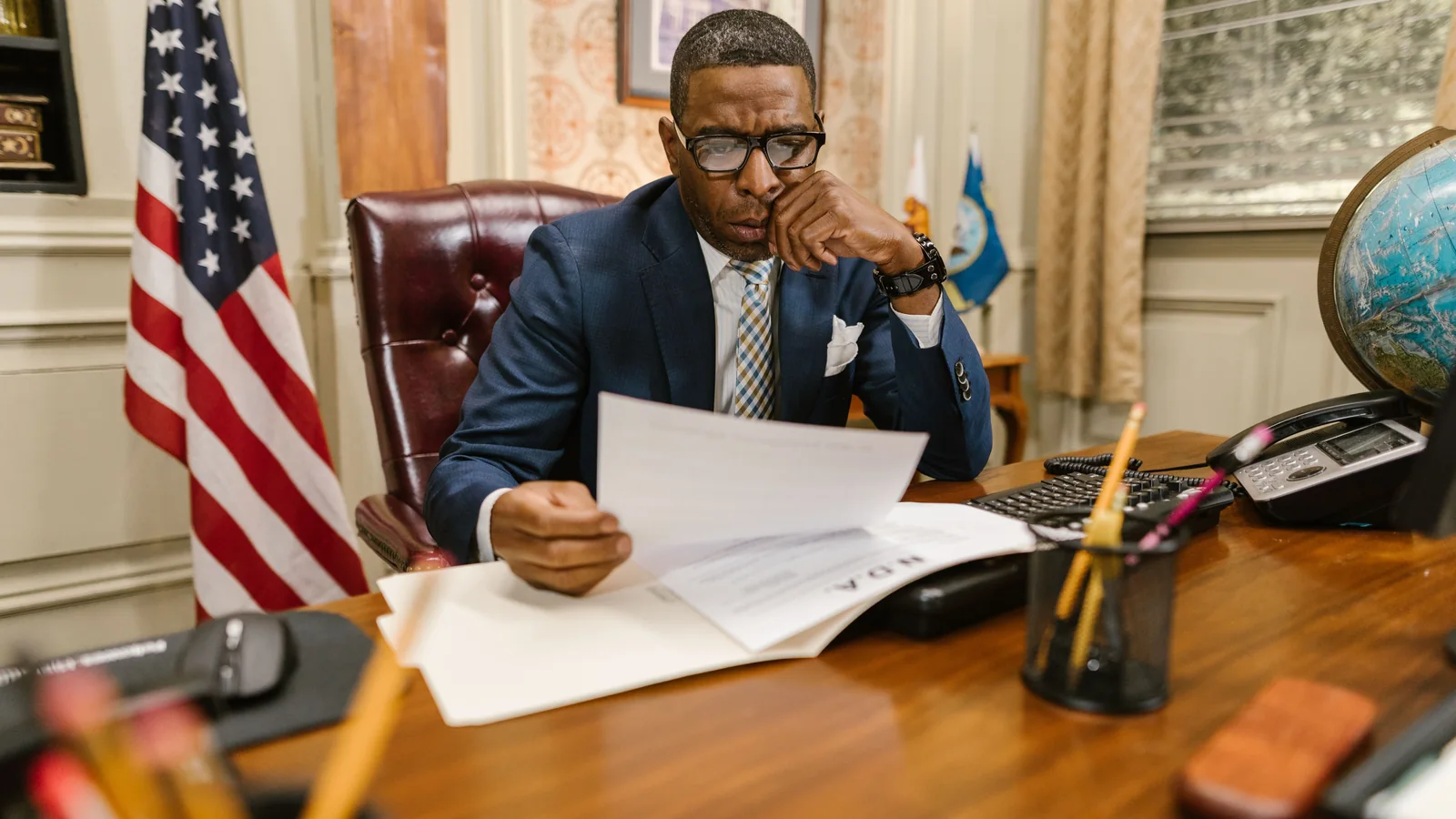 A suited professional reviews paperwork at a wooden office desk beside a U.S. flag, computer, telephone, and globe.