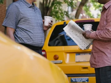 Two people stand beside a yellow car while discussing a sheet of paper; both are holding takeaway cups.