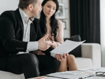 Two adults sit together at a table in a bright living room, reviewing paperwork and discussing it.