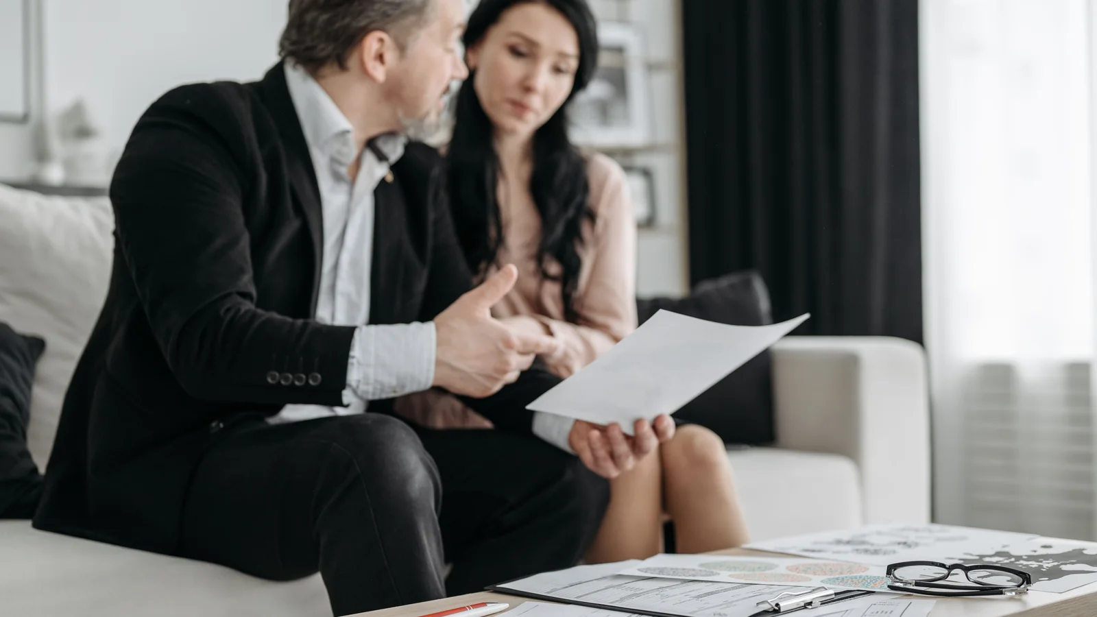 Two adults sit together at a table in a bright living room, reviewing paperwork and discussing it.