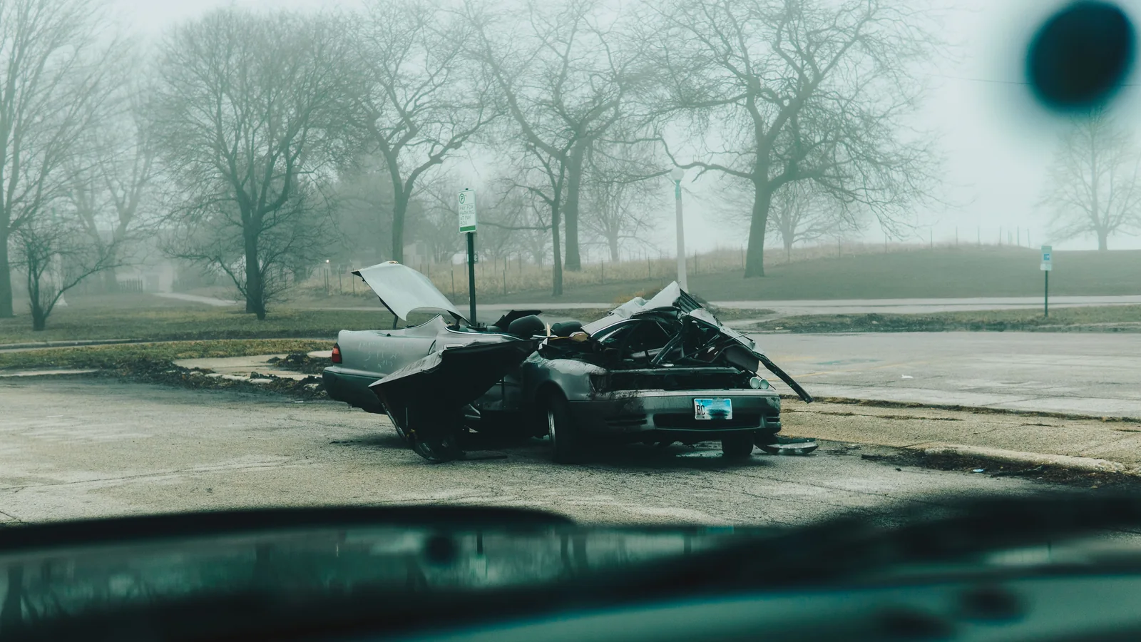 A severely damaged car sits on the side of a foggy roadside with its hood raised.
