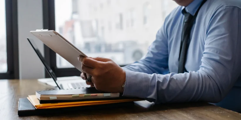 A cropped business professional in a blue shirt and tie reviews papers on a clipboard at a modern office desk.
