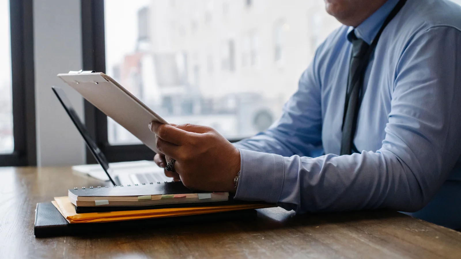 A cropped business professional in a blue shirt and tie reviews papers on a clipboard at a modern office desk.