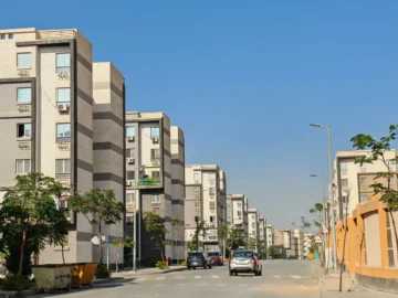 Sunlit street lined with modern apartment buildings and a few trees under a clear blue sky.