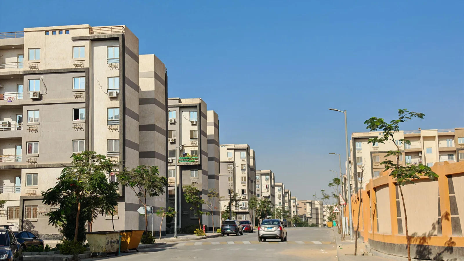 Sunlit street lined with modern apartment buildings and a few trees under a clear blue sky.