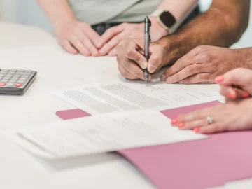 Several people review paperwork around a white table with a calculator, pens, and folders.