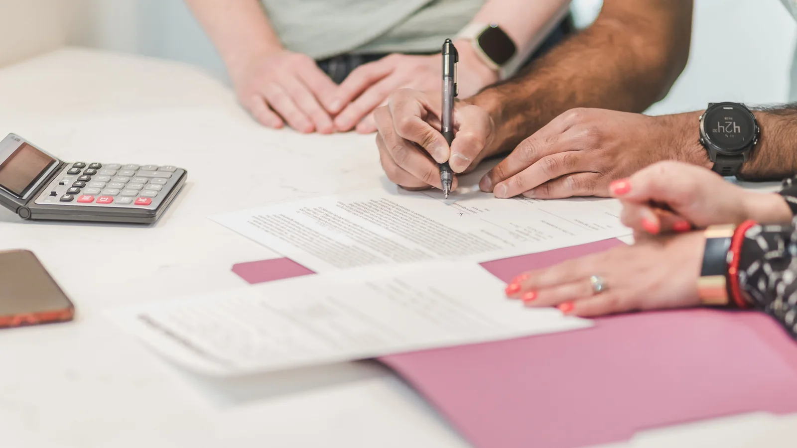 Several people review paperwork around a white table with a calculator, pens, and folders.