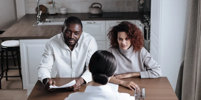 Two adults sit at a kitchen table with an advisor, discussing paperwork during a home consultation.
