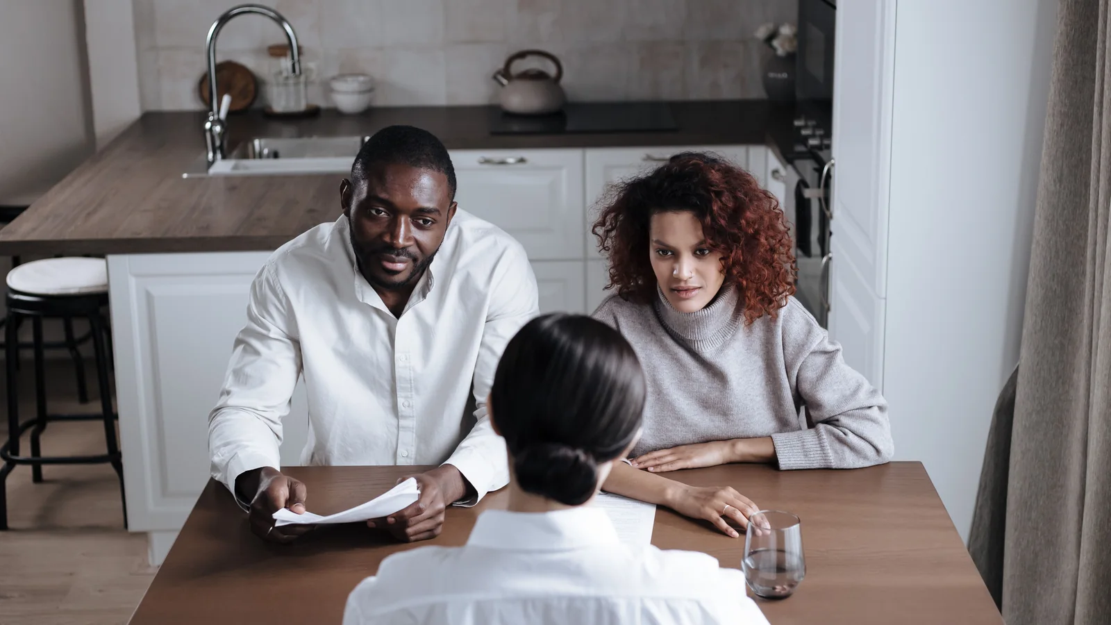 Two adults sit at a kitchen table with an advisor, discussing paperwork during a home consultation.
