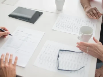 People seated around an office table reviewing paperwork, with pens, a tablet, coffee cups, and eyeglasses visible.