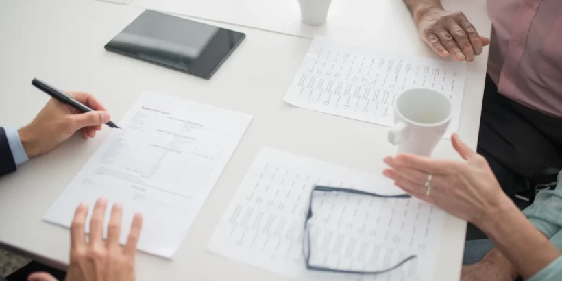 People seated around an office table reviewing paperwork, with pens, a tablet, coffee cups, and eyeglasses visible.