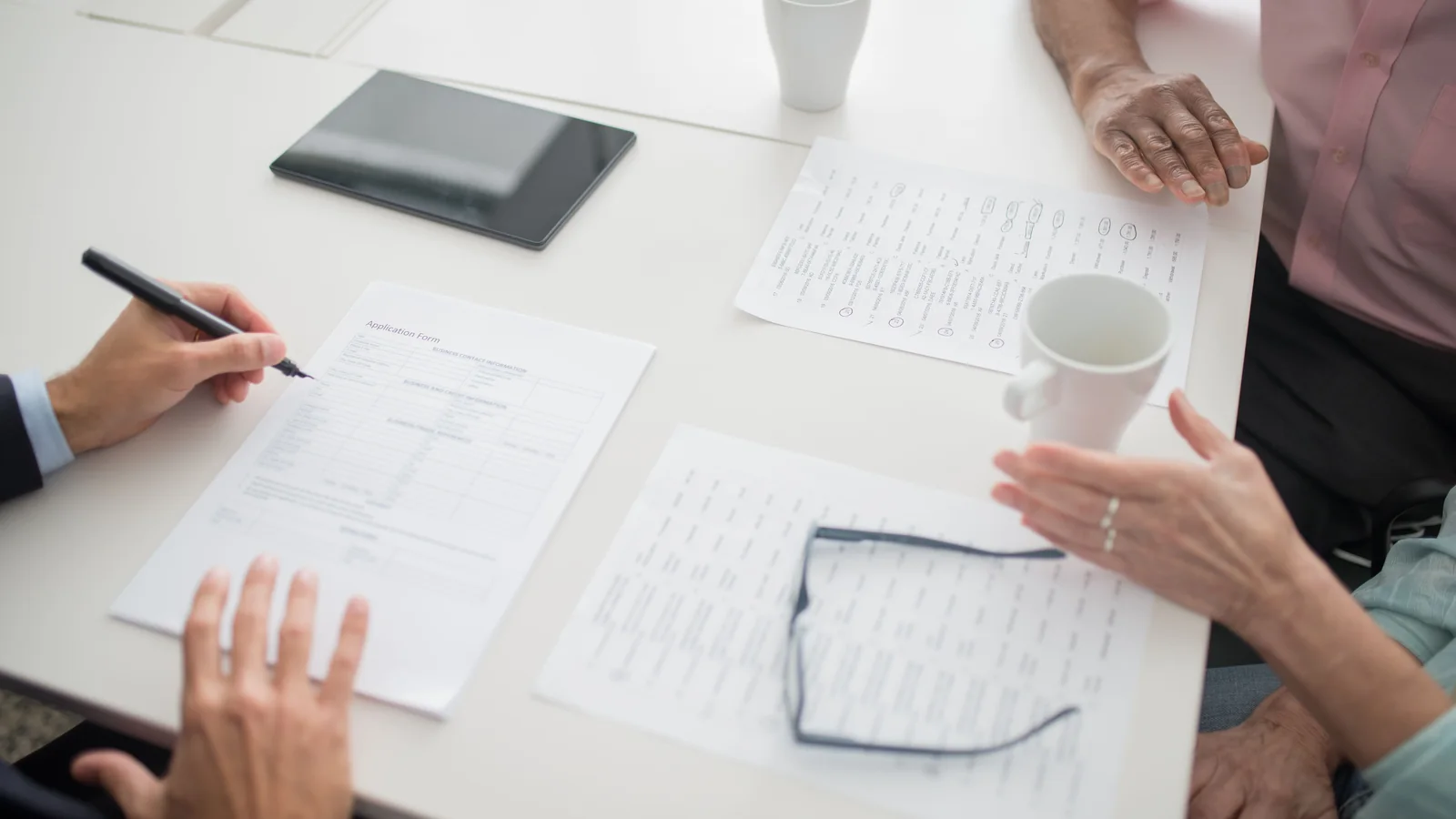 People seated around an office table reviewing paperwork, with pens, a tablet, coffee cups, and eyeglasses visible.