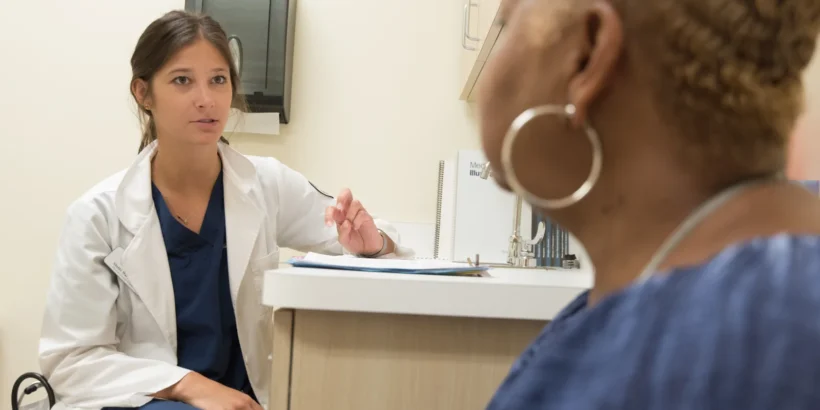 Doctor in a white coat speaks with a seated patient in a medical examination room.