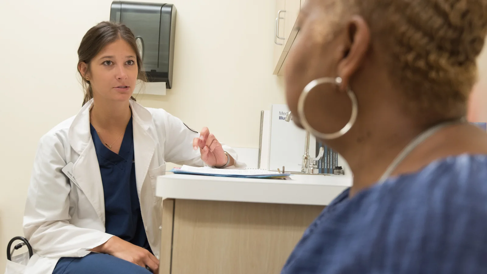 Doctor in a white coat speaks with a seated patient in a medical examination room.