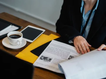 Overhead view of a professional reviewing printed documents at a dark office table beside a tablet, coffee cup, and yellow folder.