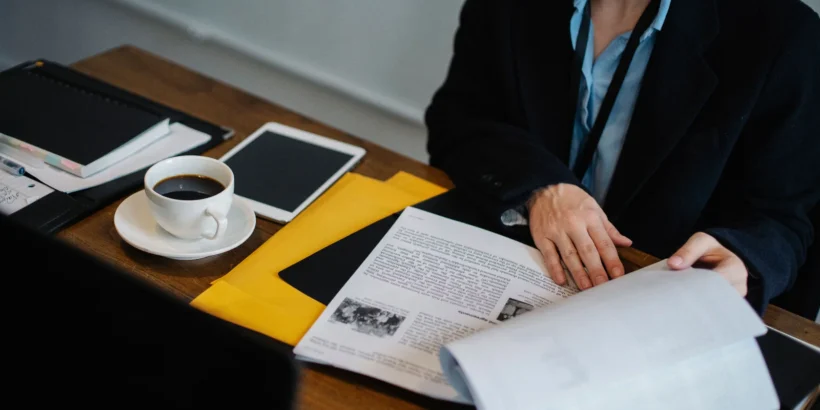 Overhead view of a professional reviewing printed documents at a dark office table beside a tablet, coffee cup, and yellow folder.