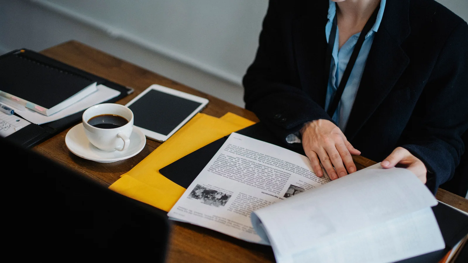 Overhead view of a professional reviewing printed documents at a dark office table beside a tablet, coffee cup, and yellow folder.