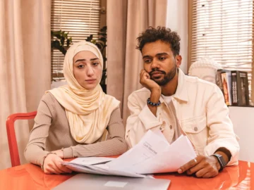 A couple sits at a table reviewing paperwork beside a laptop in a bright home office.