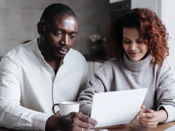 A couple sits together at a kitchen table reviewing paperwork beside coffee cups.