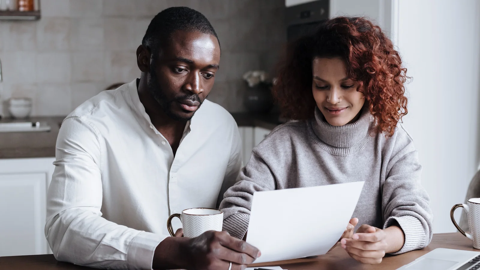 A couple sits together at a kitchen table reviewing paperwork beside coffee cups.