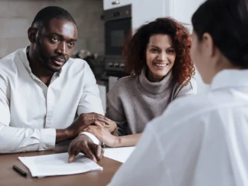 A couple sits at a table with a professional advisor, reviewing paperwork together in a home setting.