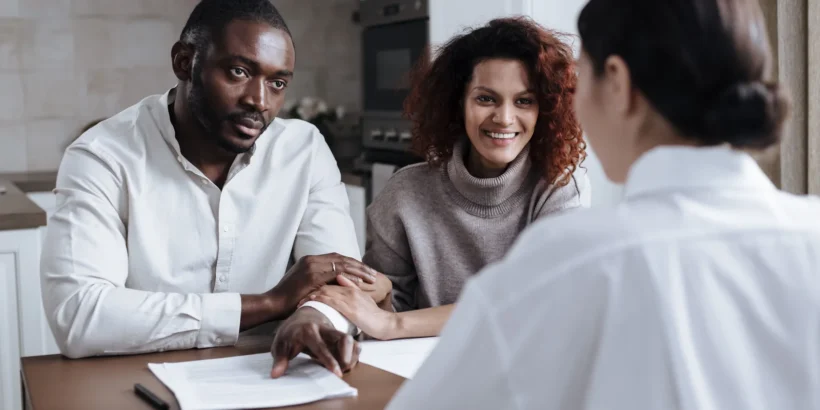 A couple sits at a table with a professional advisor, reviewing paperwork together in a home setting.