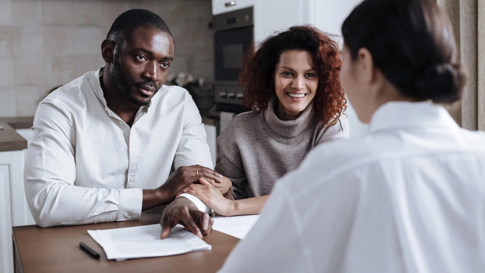 A couple sits at a table with a professional advisor, reviewing paperwork together in a home setting.