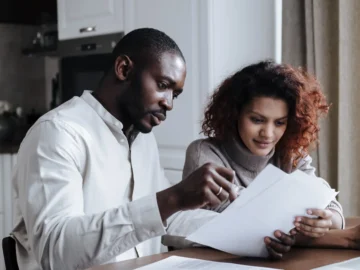 A couple sits at a kitchen table reviewing documents together beside a laptop and coffee.