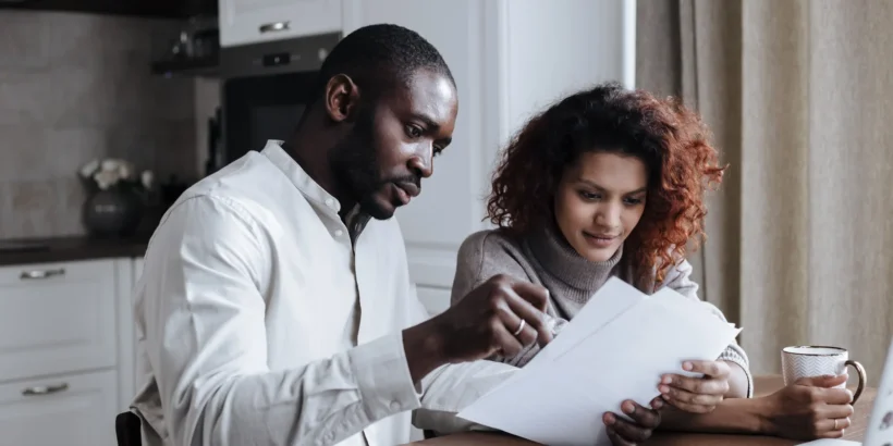 A couple sits at a kitchen table reviewing documents together beside a laptop and coffee.