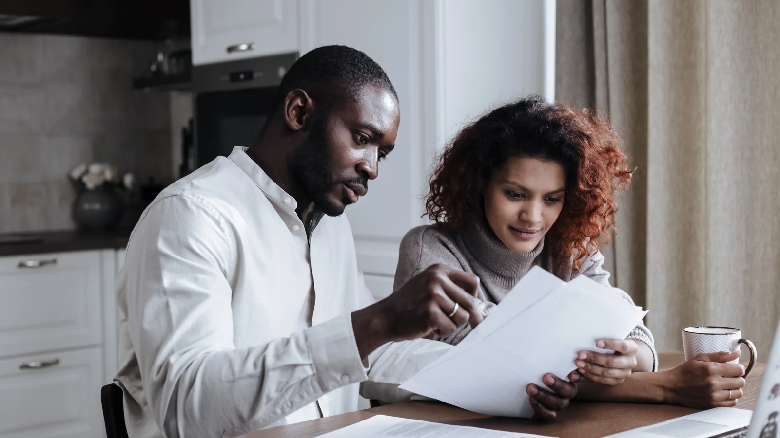 A couple sits at a kitchen table reviewing documents together beside a laptop and coffee.